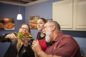 Three people in a kitchen admiring a huge BLT, one of whom is getting ready to take a huge bite of it. 