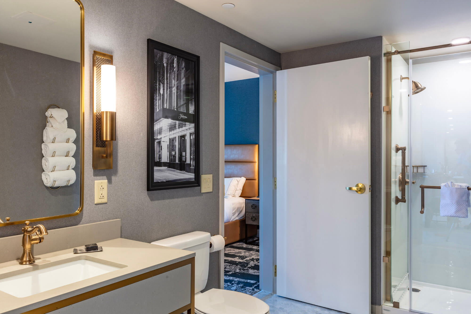 A hotel bathroom with the sink and toilet in the foreground, shower and slight view through tot he bedroom in the background.