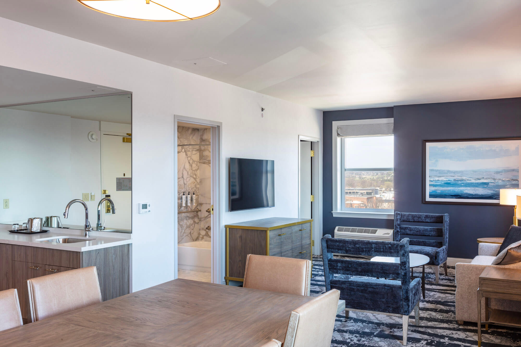 A hotel suite with a dinning table in the foreground and a seated lounge area with TV and sink in the background.