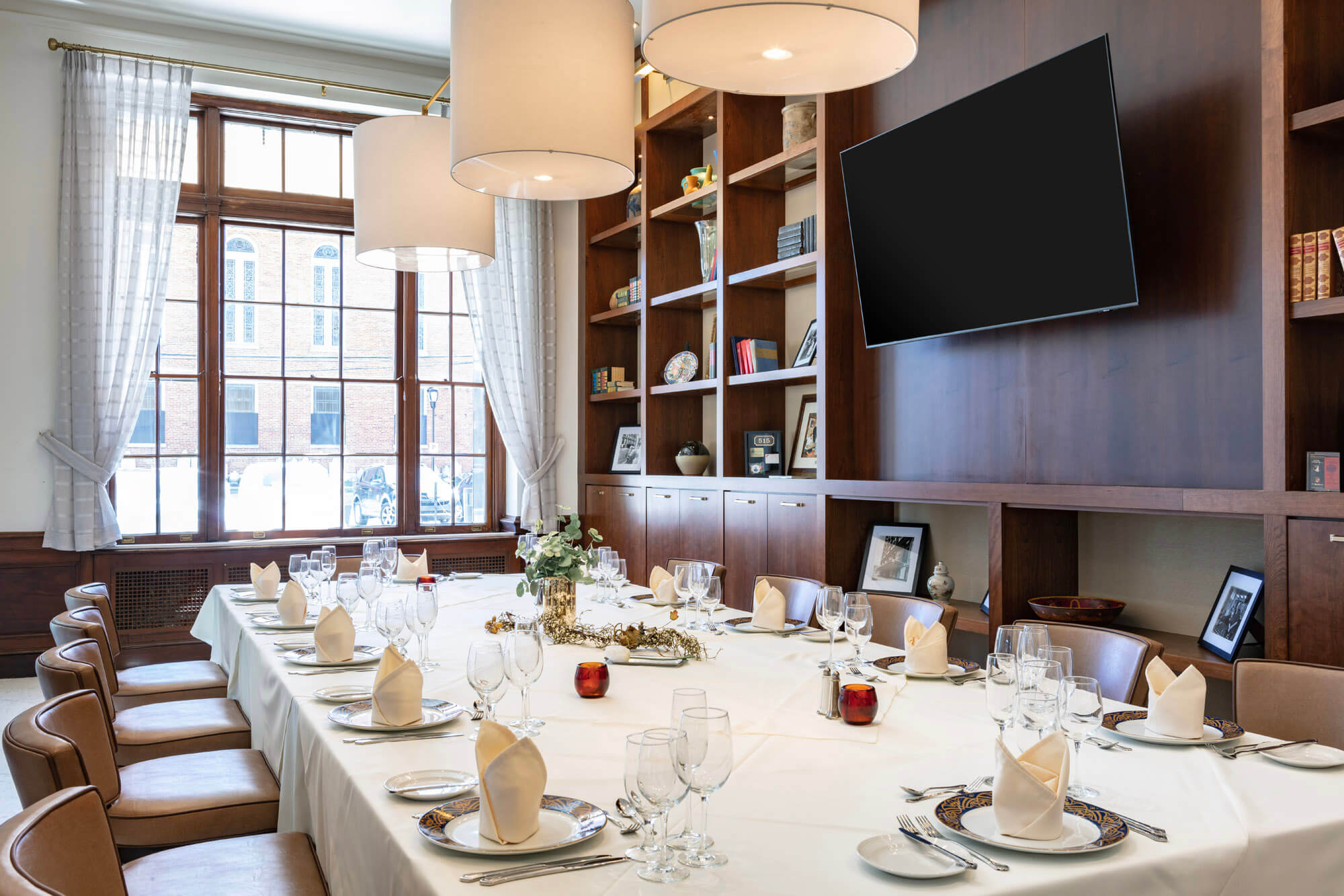 Large table with dinning set up for twelve, a large tv on the wall and a window in the background looking to the street.