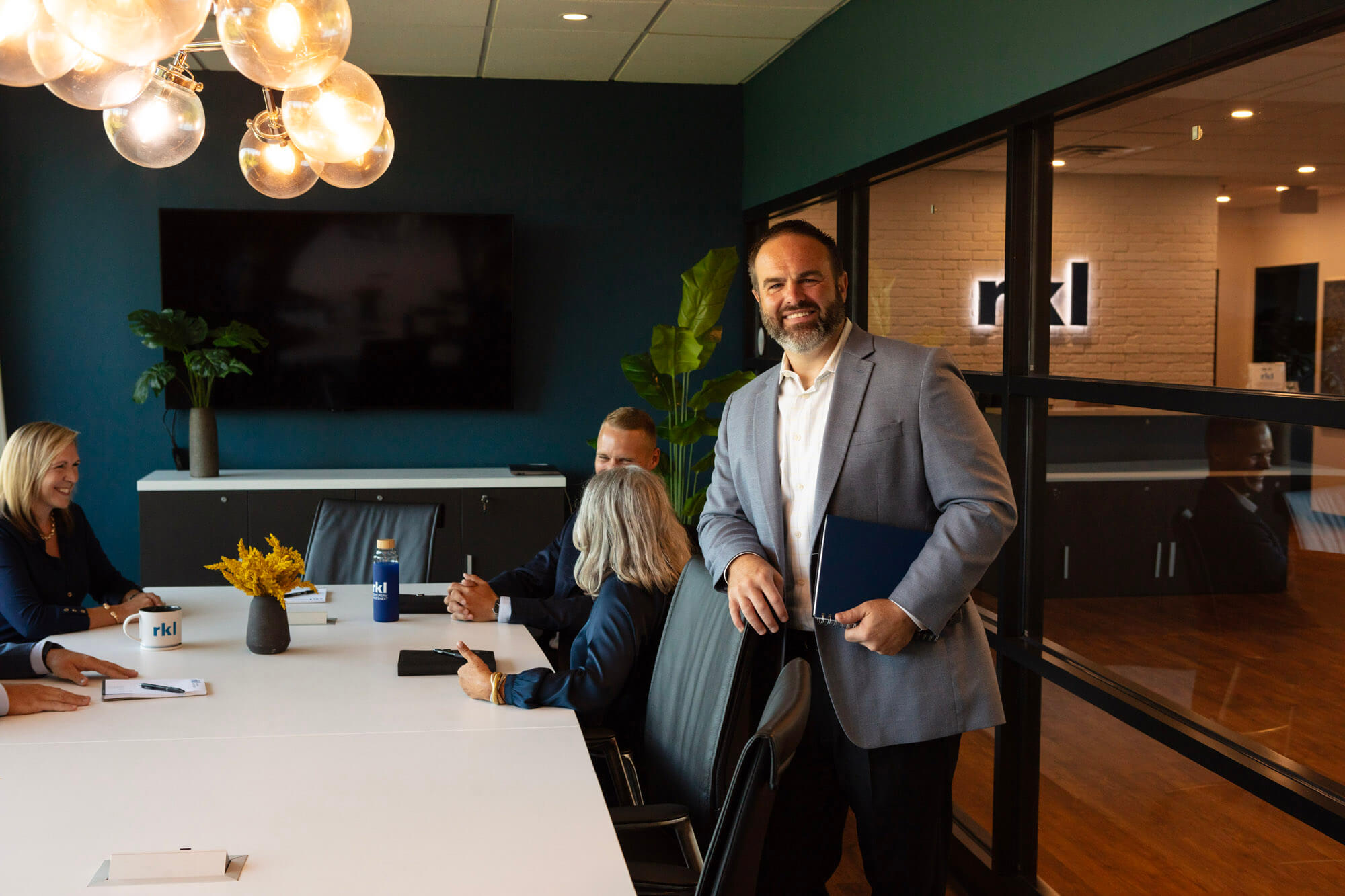 A man in the foreground smiling at the camera, colleagues in the backgrounds talking around a conference table.