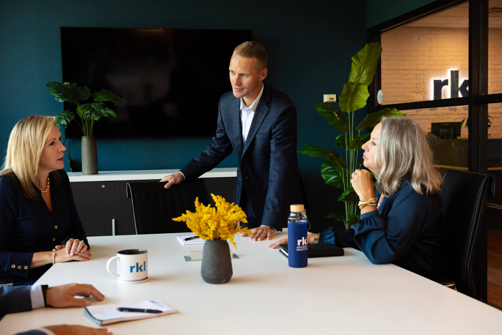 A man standing and two women sitting around a conference table talking.