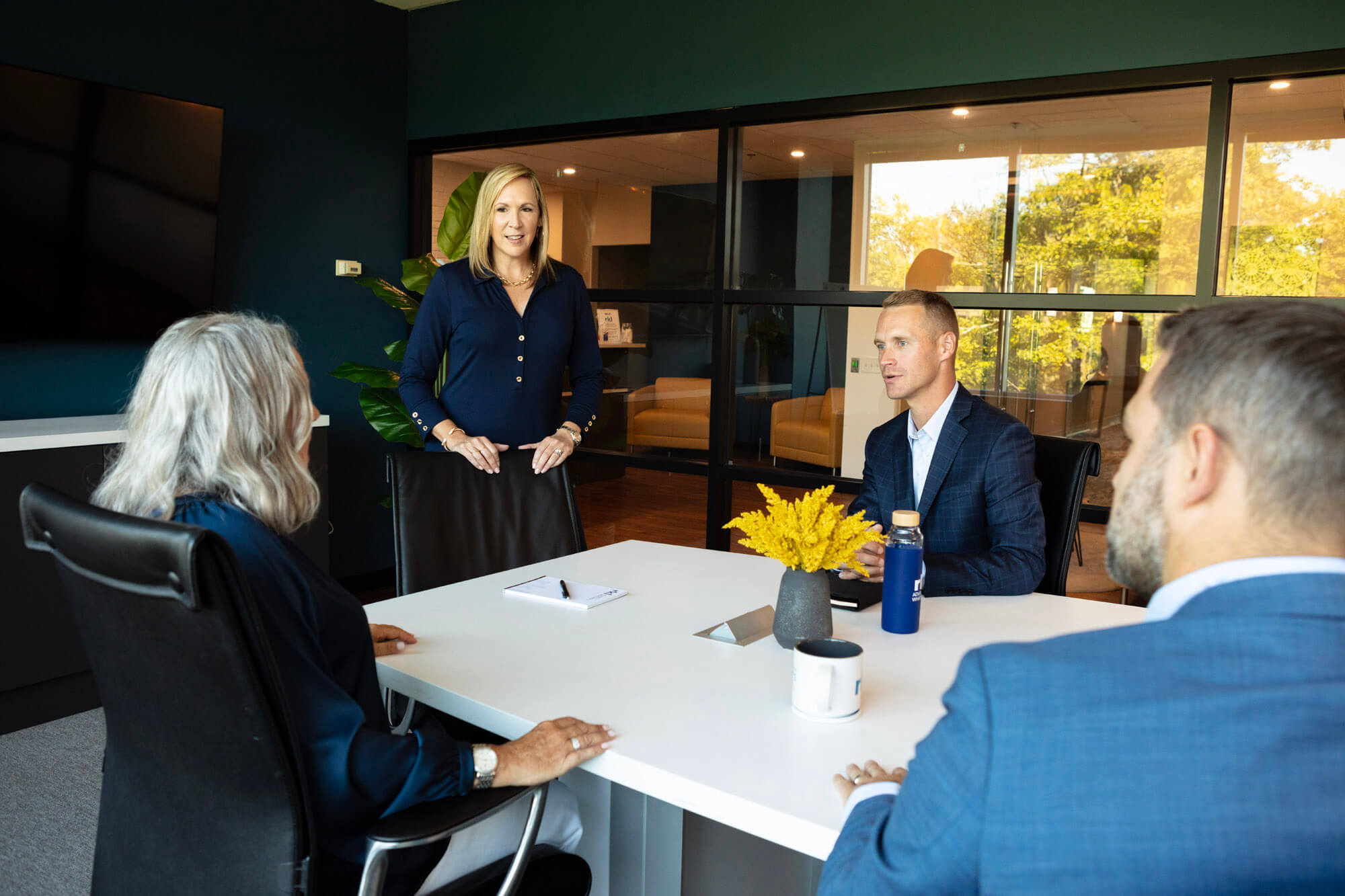 A woman standing and three other people sitting and talking around a conference table.