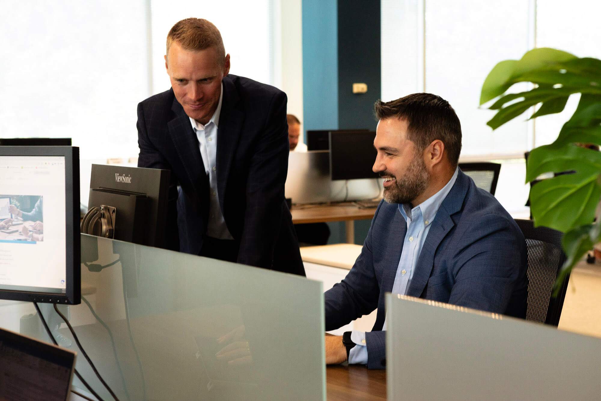 a man sitting at a desktop computer and talking to another man who is looking at the monitor.