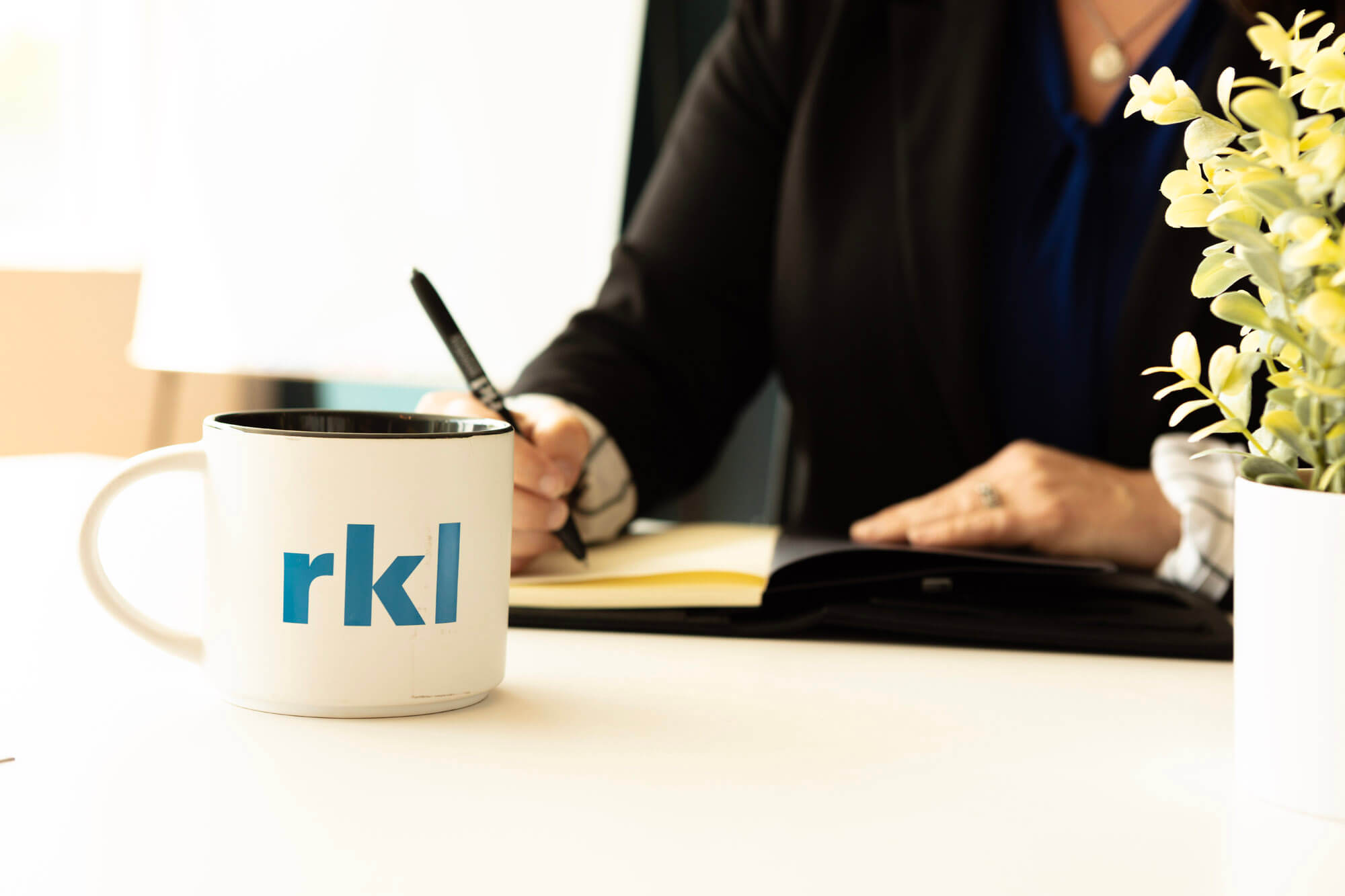 A RKL branded coffee mug in the foreground and a woman in the background writing notes at a table.