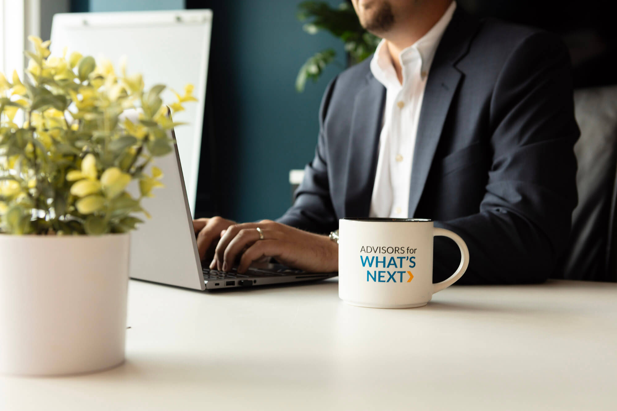 A RKL branded coffee mug in the foreground and a man in the background typing on his laptop at a table.