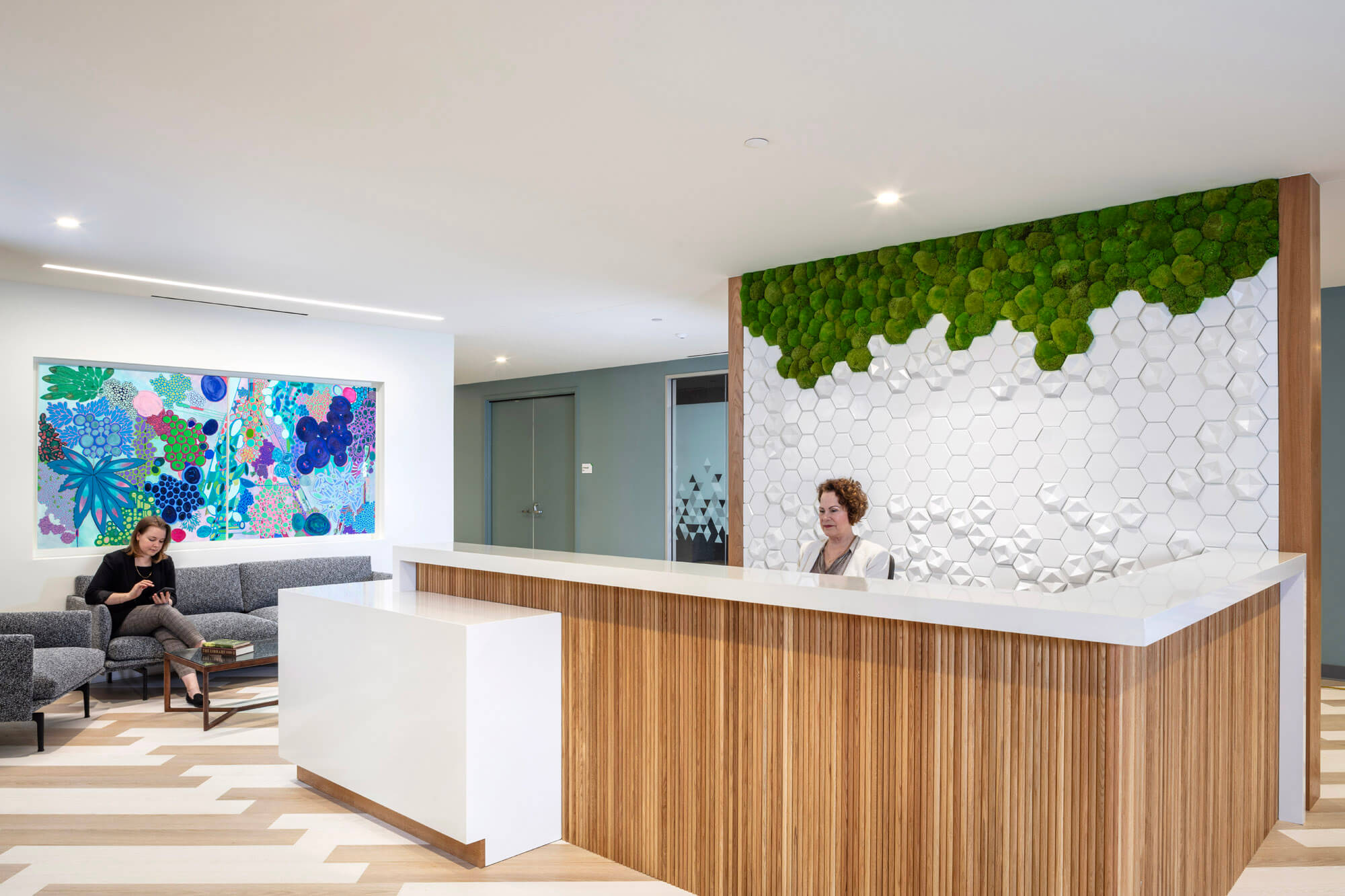 front desk of a commercial space with a lady sitting behind the desk and a woman waiting on a sofa to the side.