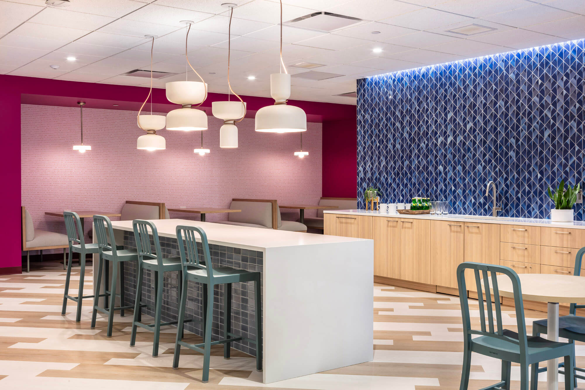 Bright kitchen area in a commercial office with an island bench and blue tile splash back and pink booths on the back wall.