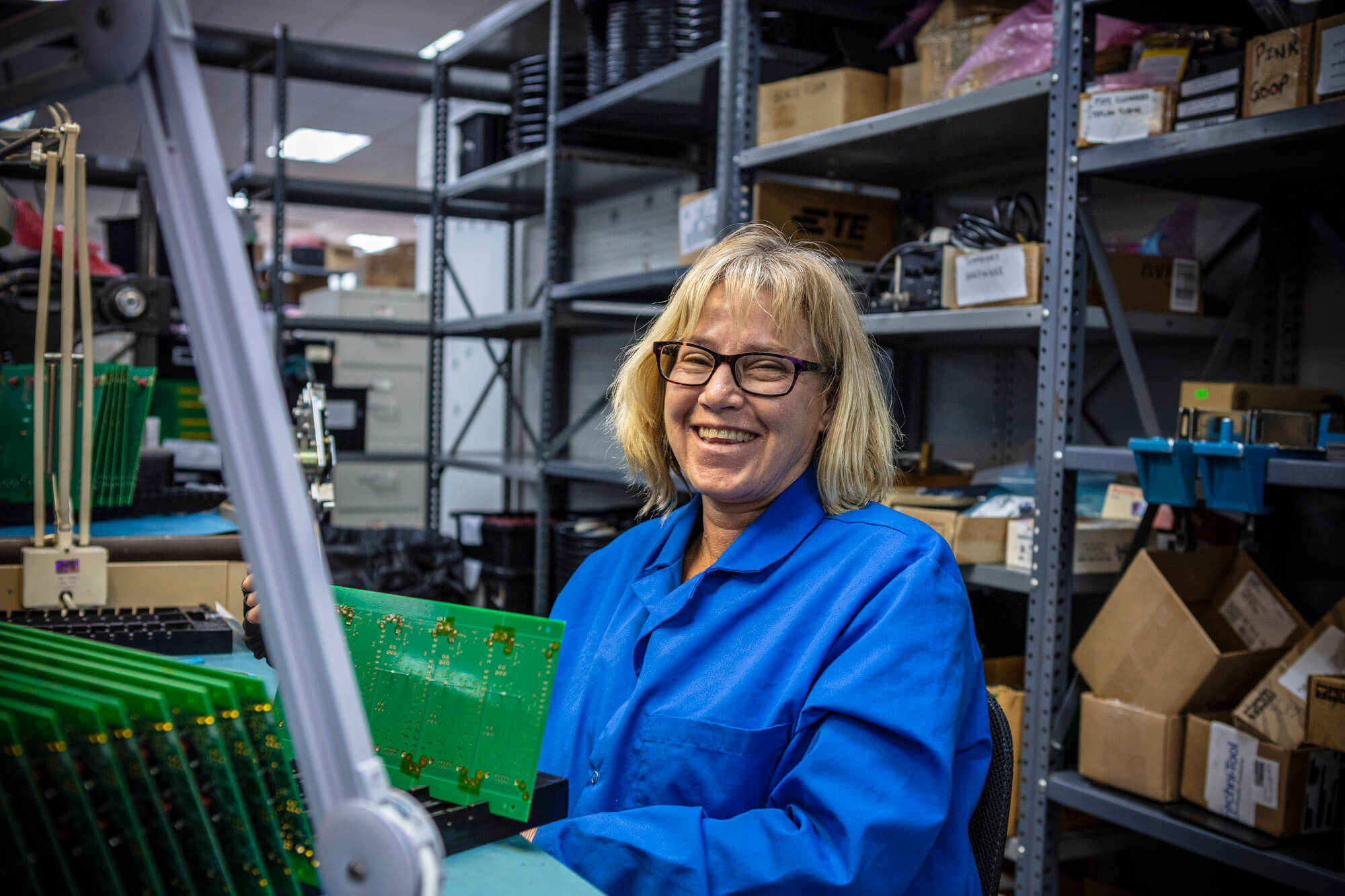 A lady laughing with a blue coat on and she is working on a manufacturing a green circuit board.