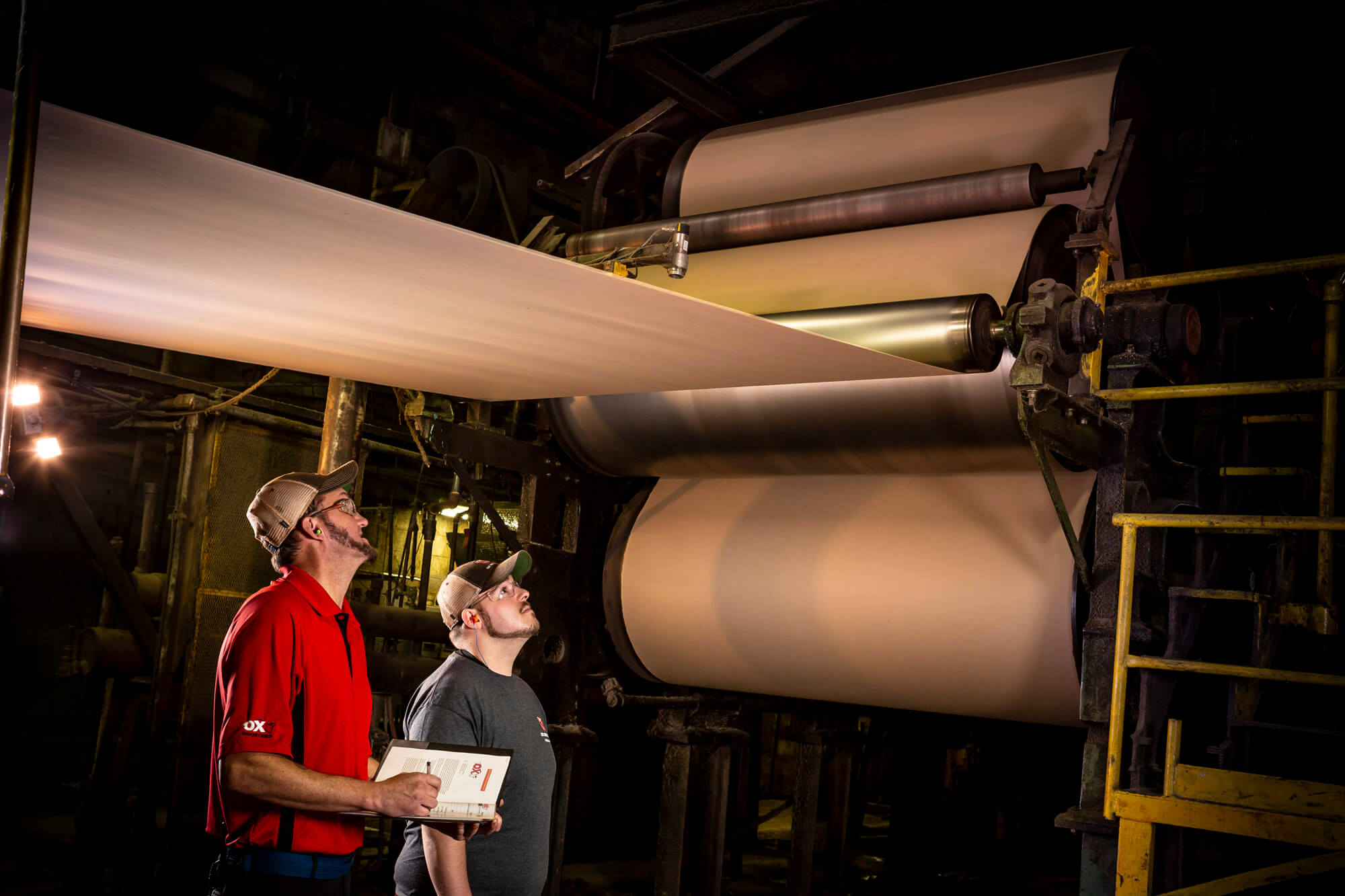 Two men standing under a huge roll of paper in a factory.