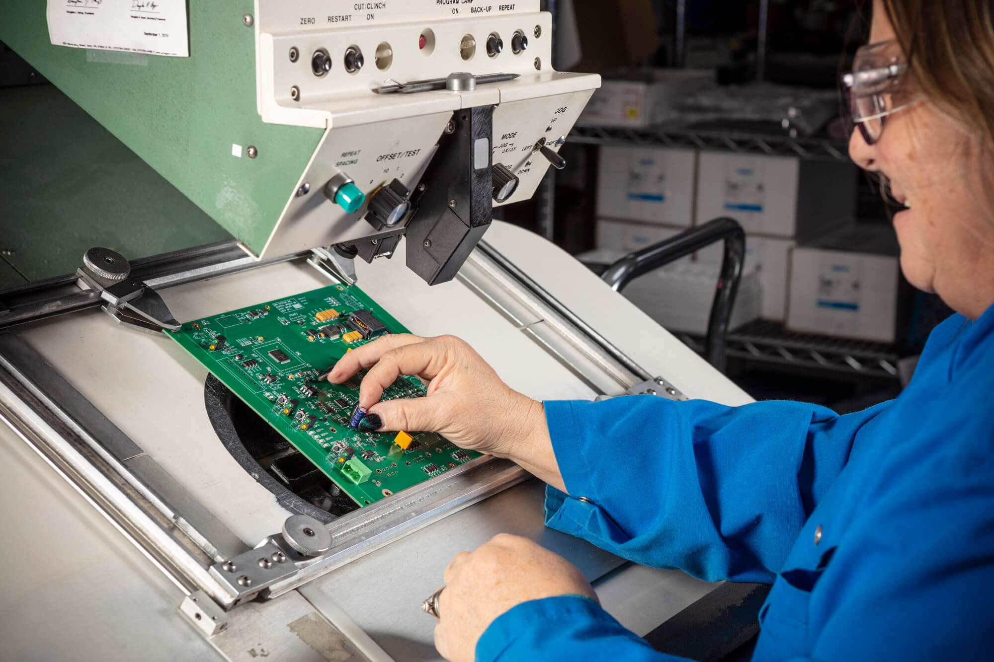 Woman in blue coat working closely on a green circuit board.