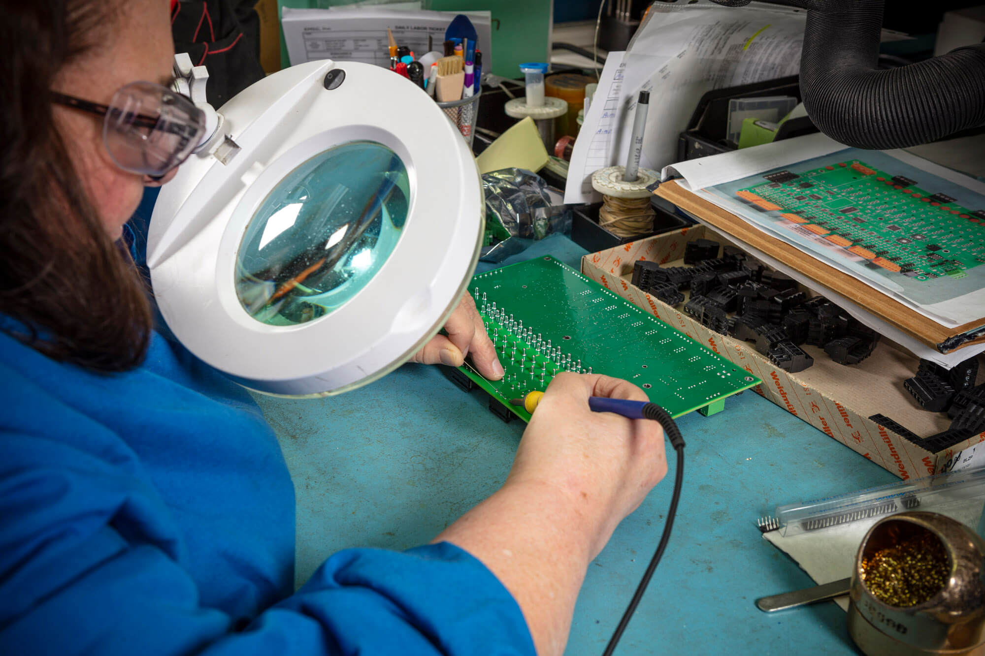 A woman in a blue coat looking through a magnifying glass creating with a soldering iron on a circuit board.