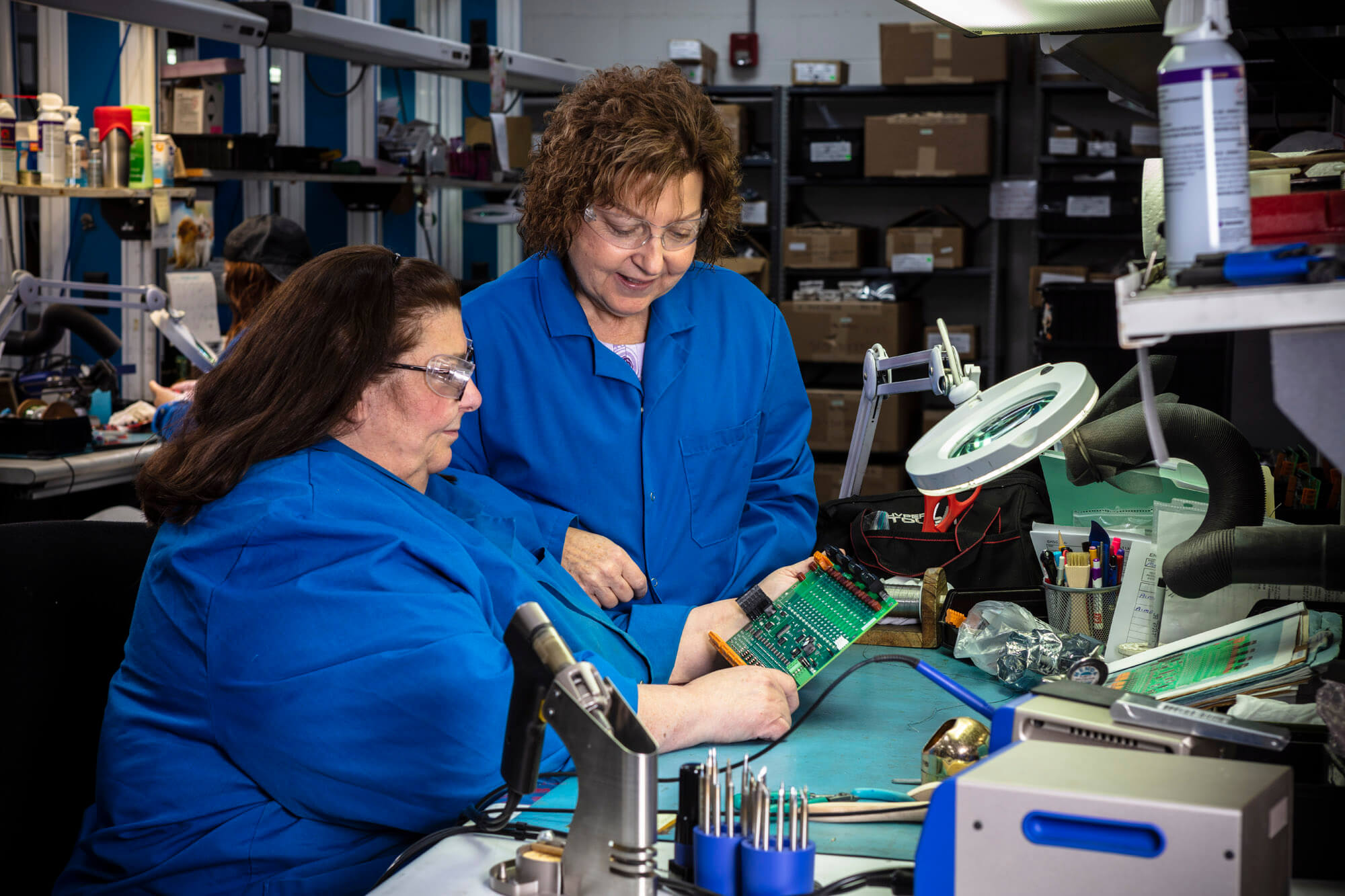 Two women in blue coats looking at a green circuit board.