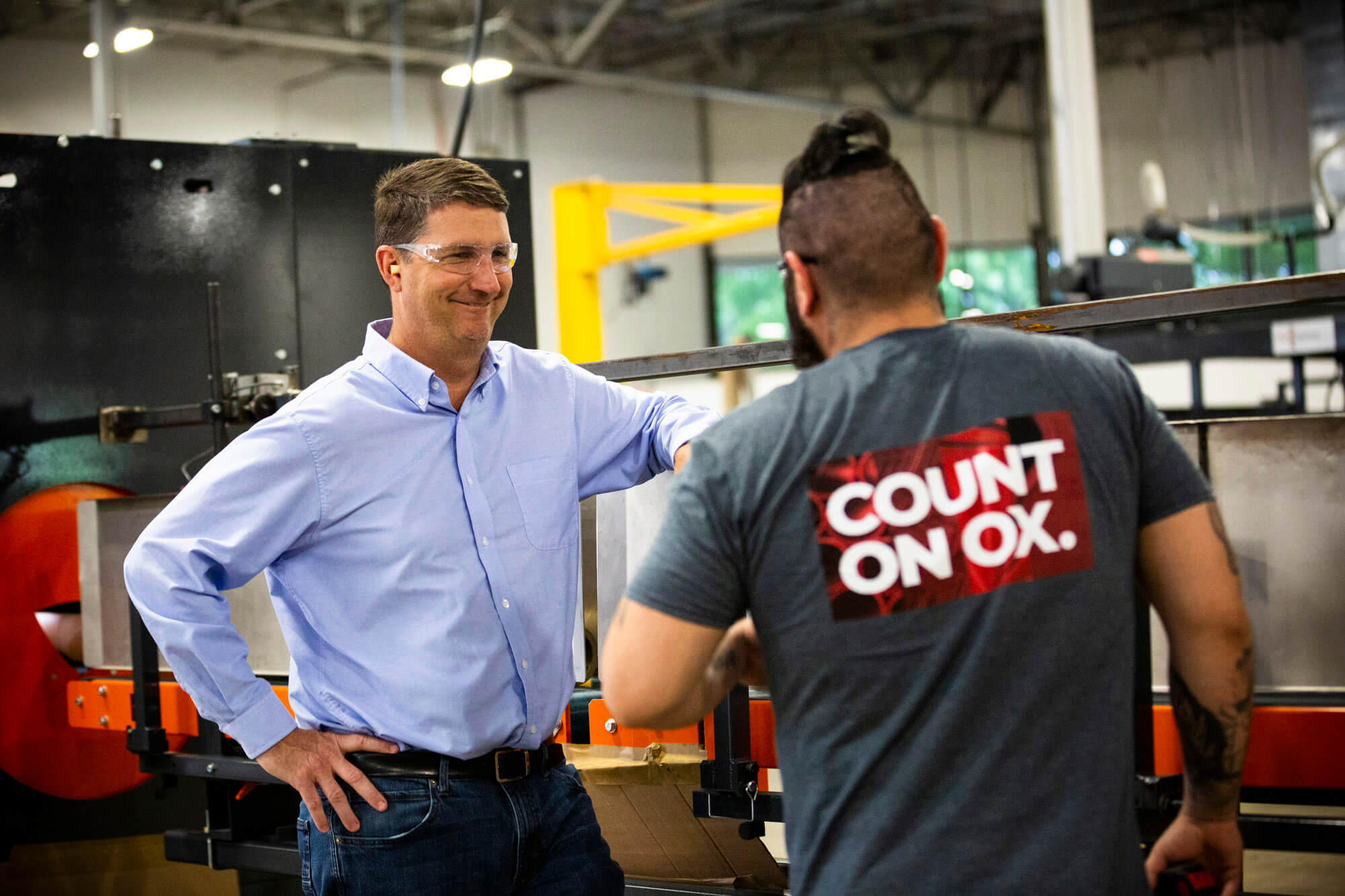 Two men, one in an ox industries t-shirt, the other in a business shirt. They are talking in a factory.