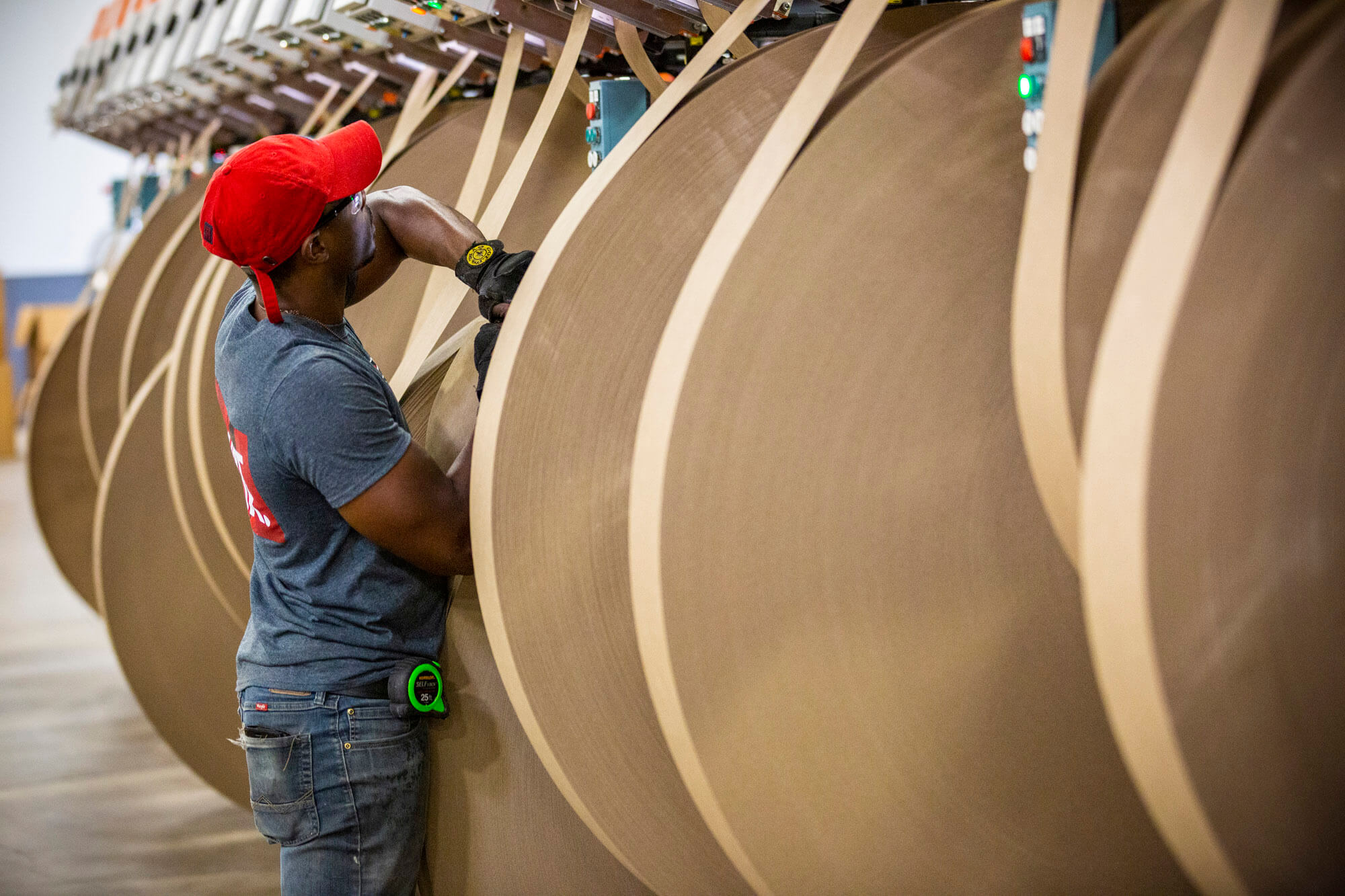 A man with a red hat on working with a large roll of cardboard in a factory.