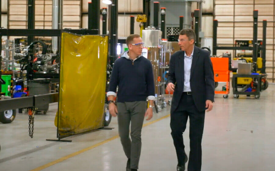 Two men walking towards the camera looking at each other talking. They are on a factory floor.