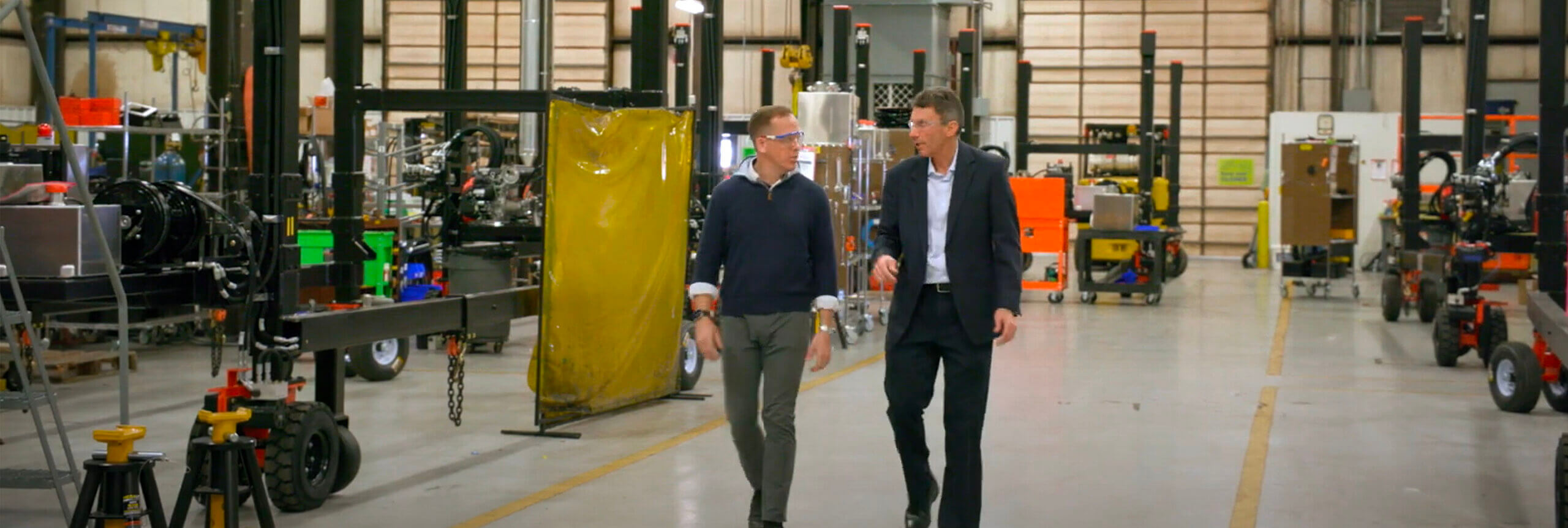 Two men walking towards the camera looking at each other talking. They are on a factory floor.