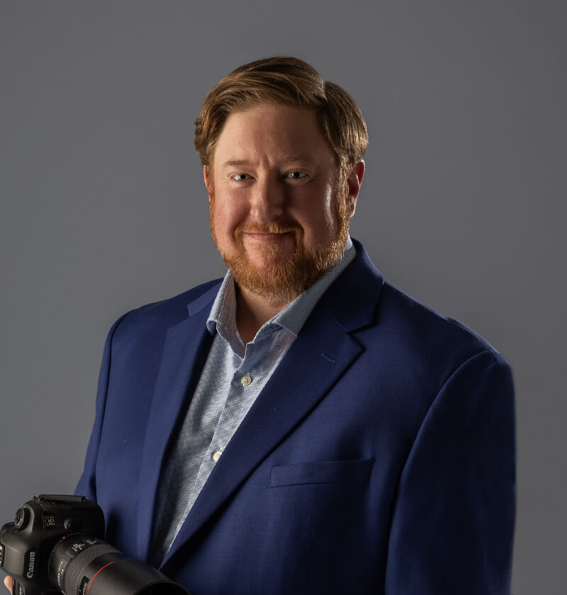 Man standing looking at the camera with a sports jacket on and a camera in his hand.