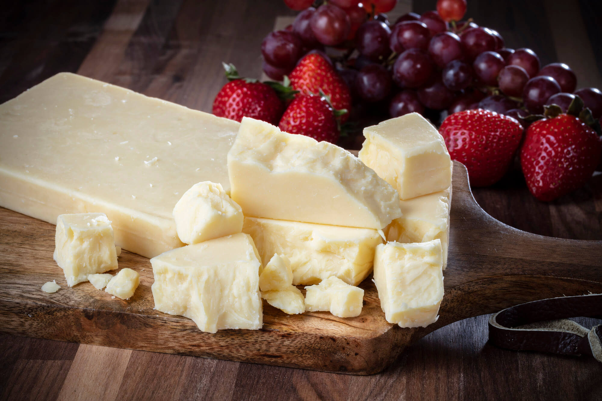 A block of cheese broken up and laying on a wooden board with strawberries and grapes in the background.