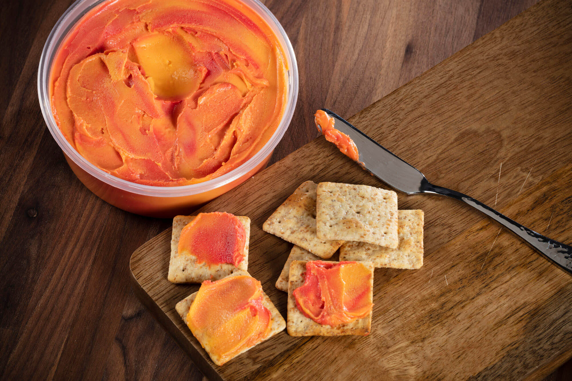 A top down shot of a container of cheese spread and crackers next to it with spread on a wooden board.