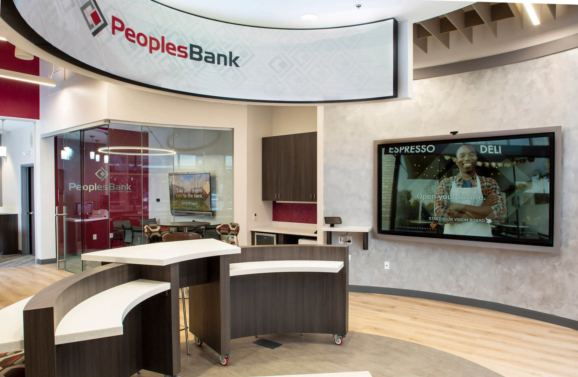 Image of the branch of Peoples Bank with a desk, office , and large TV screen in the back ground.