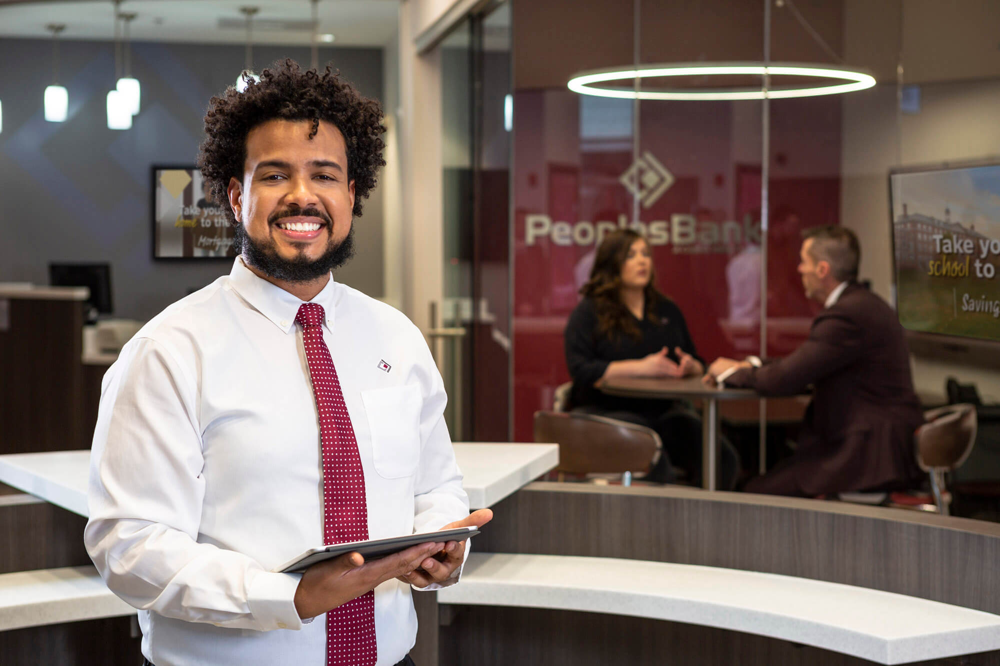 A man holding an iPad in the foreground and two people talking in a glass office in the background.