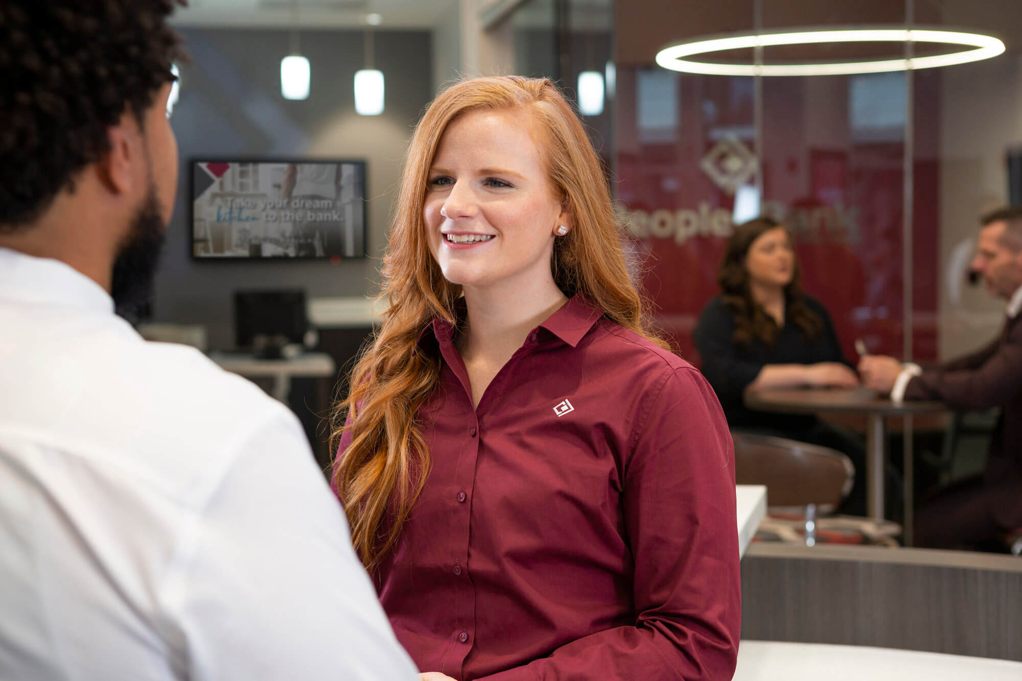 A man talking to a woman in the foreground with a blurred man and woman talking in a glass office in the background.