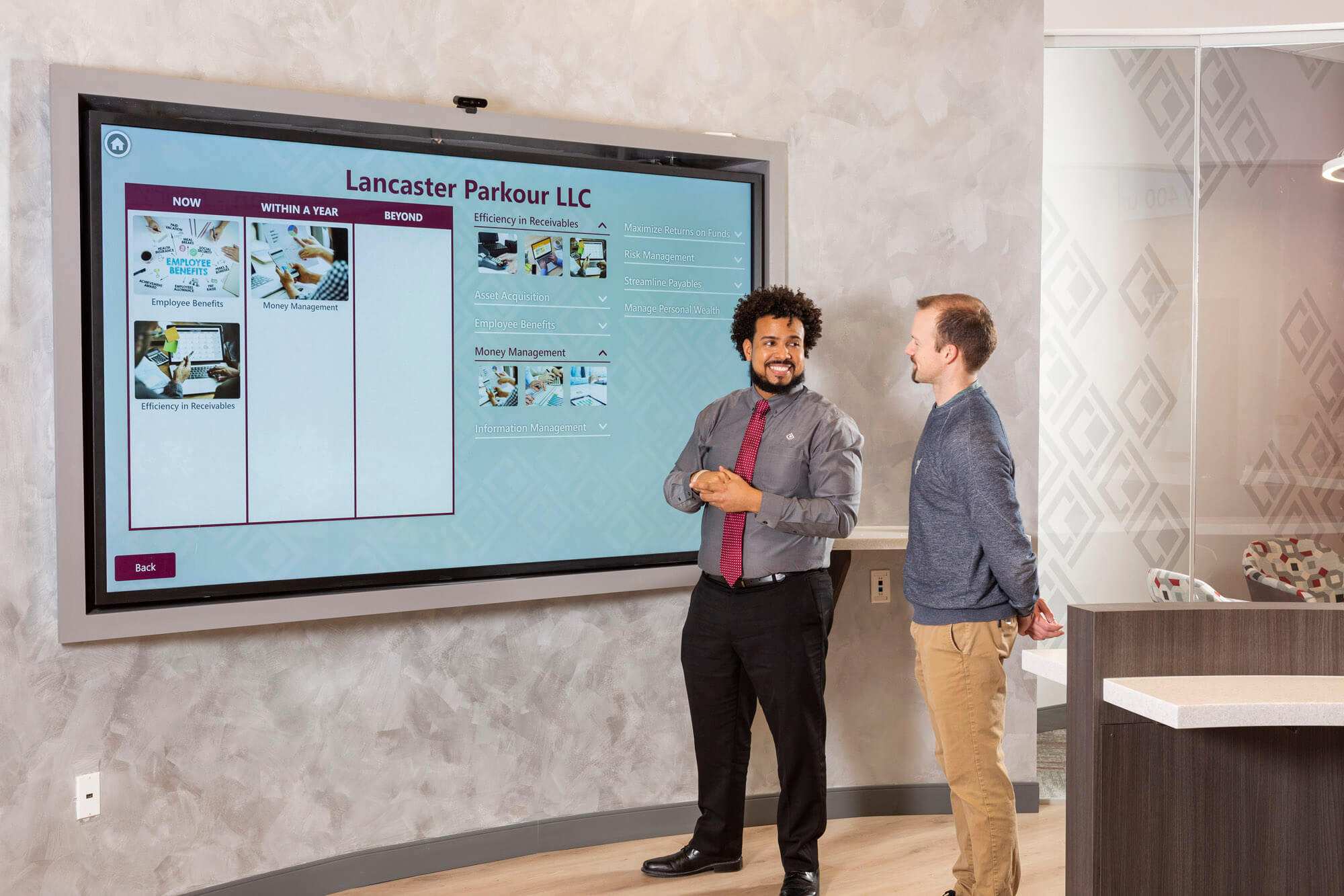 A man smiling at another man in front of a large interactive screen.