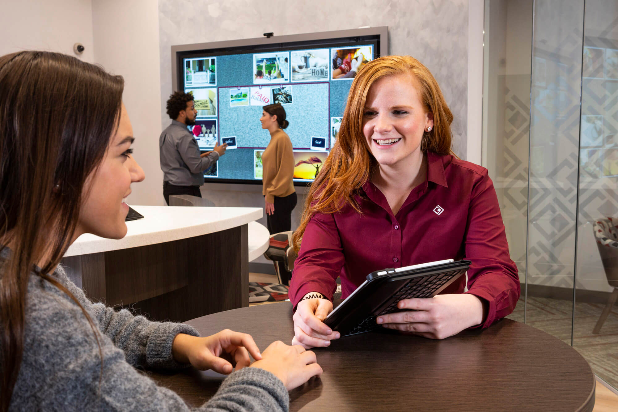 Two people in the foreground smiling and looking at an iPad on a desk. In the background there is a man and woman looking at a large screen.