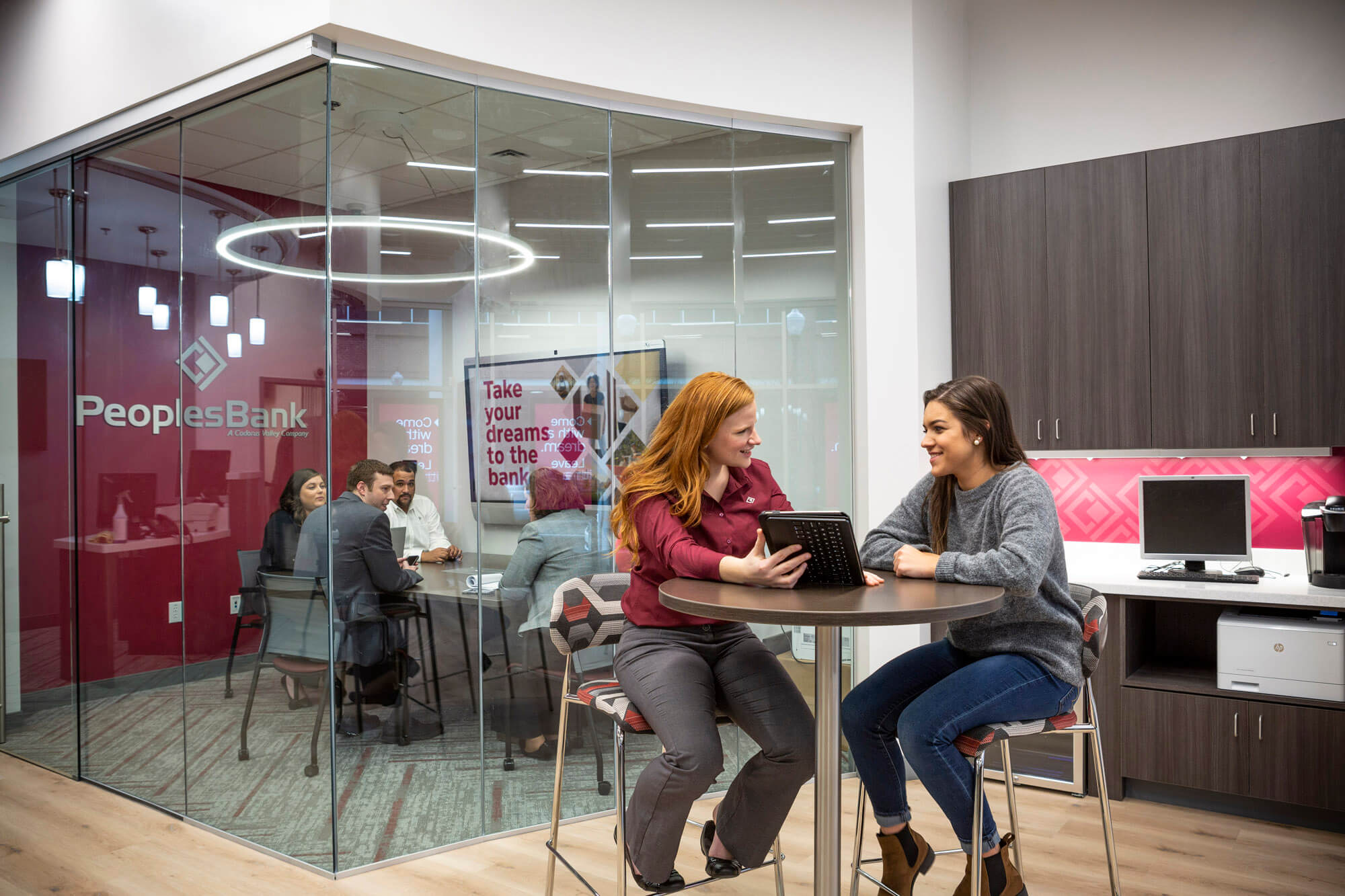 Two people sitting at a high top table looking at an iPad while a group of four people are meeting in a glass office in the background.