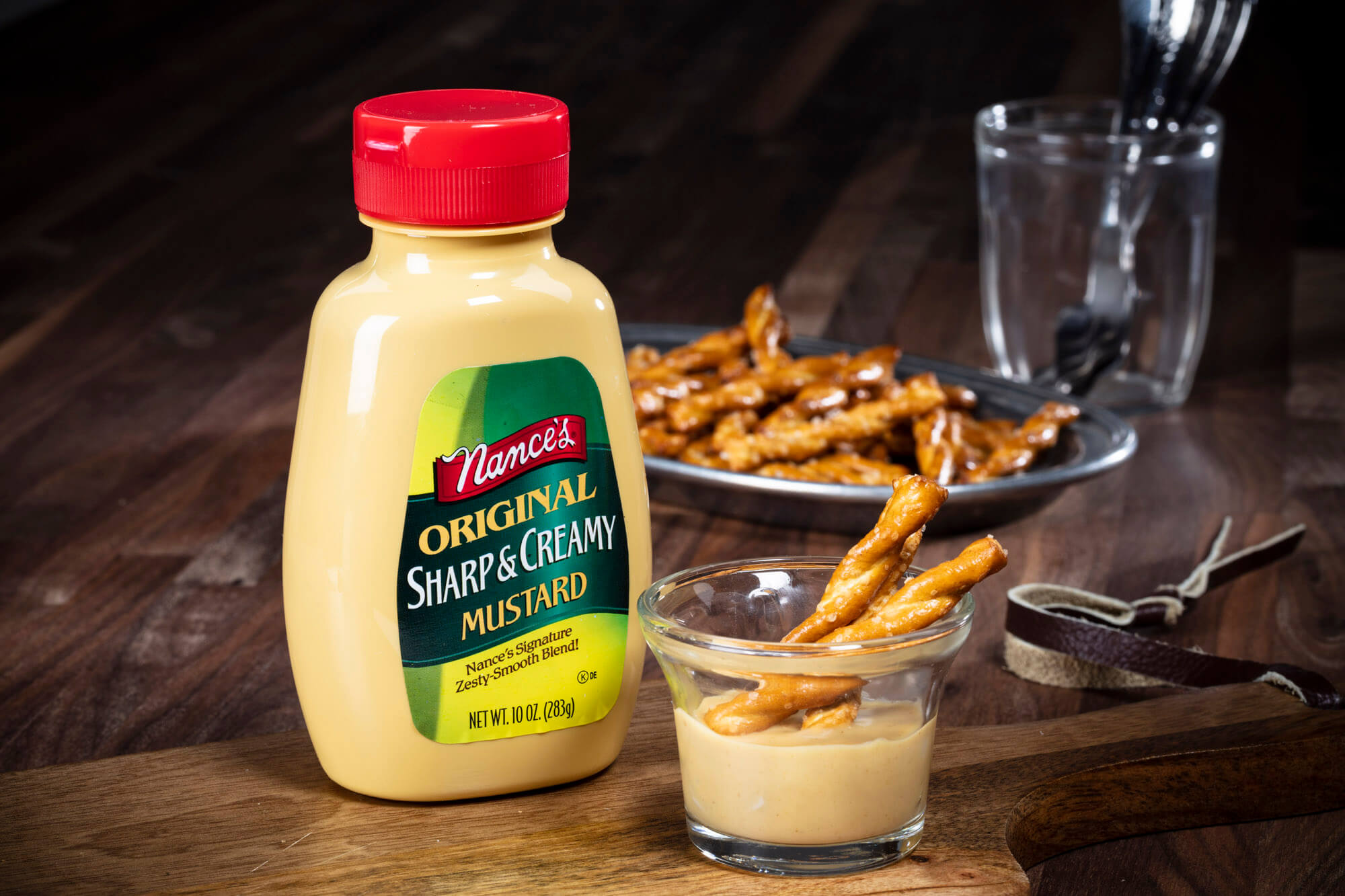 Container of mustard with a small glass cup and pretzel sticks.