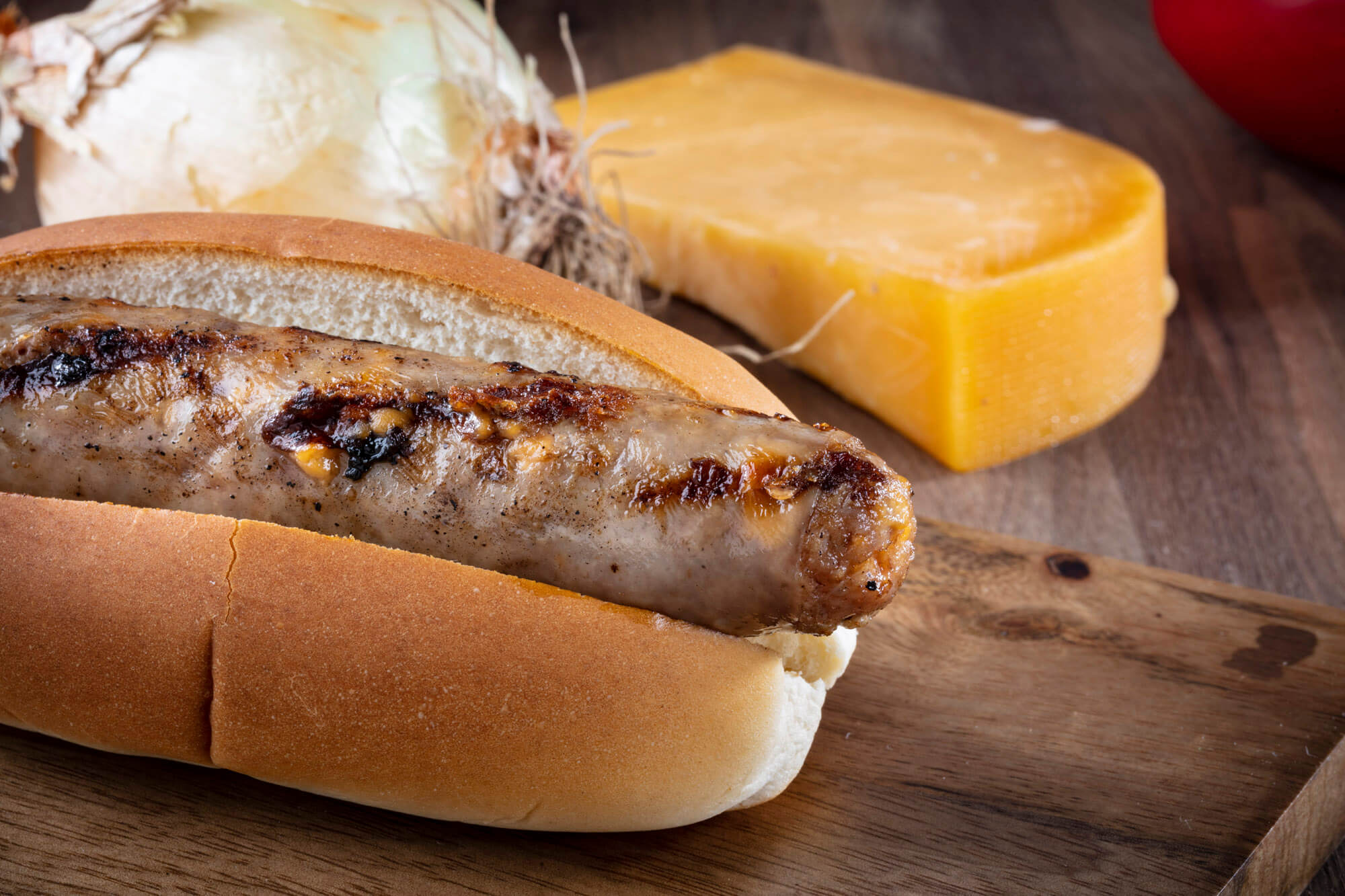 A sausage in a bread roll on a wooden board with a block of cheese in the background.