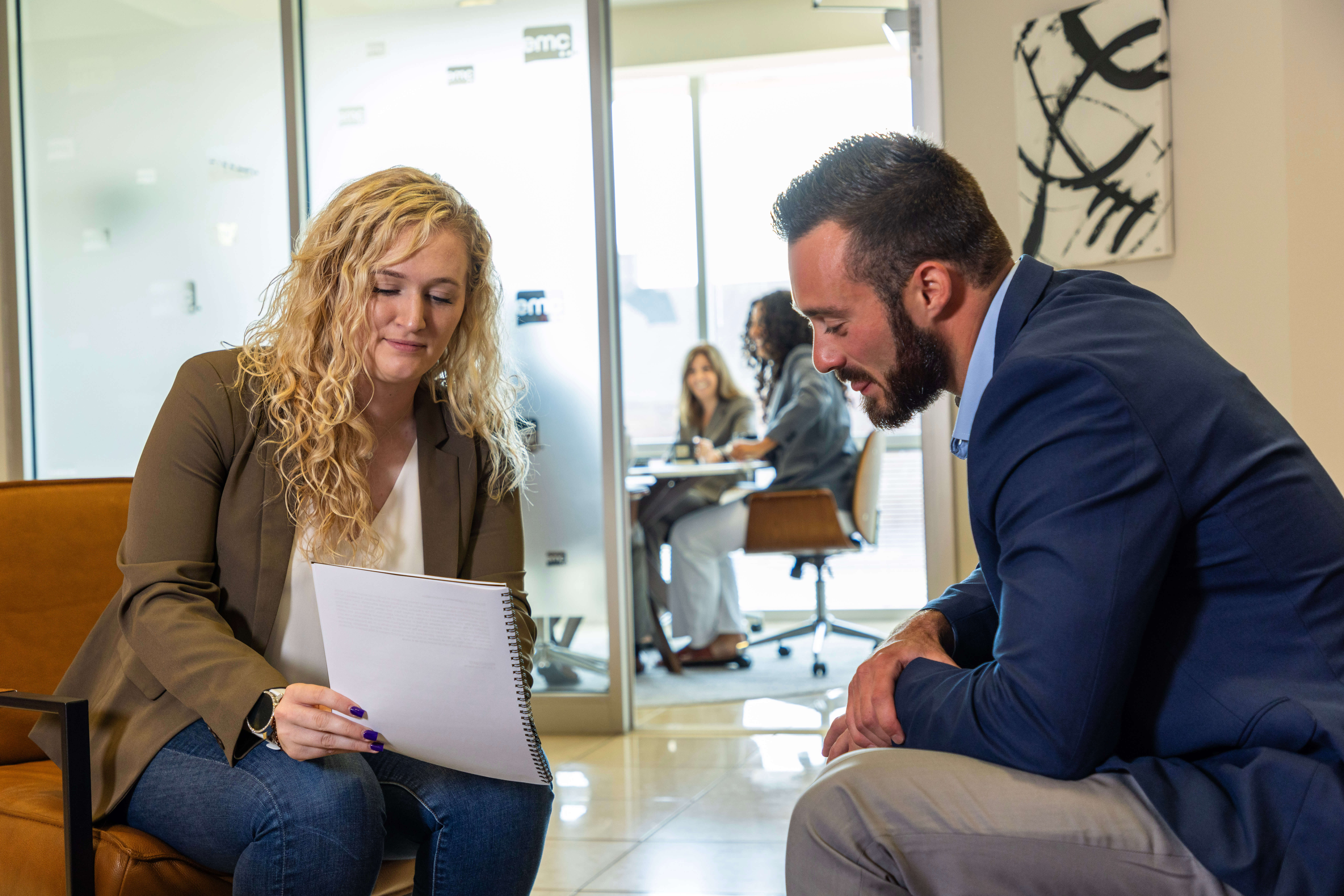 Two people in a professional setting looking at a document. There are people in an office behind them.